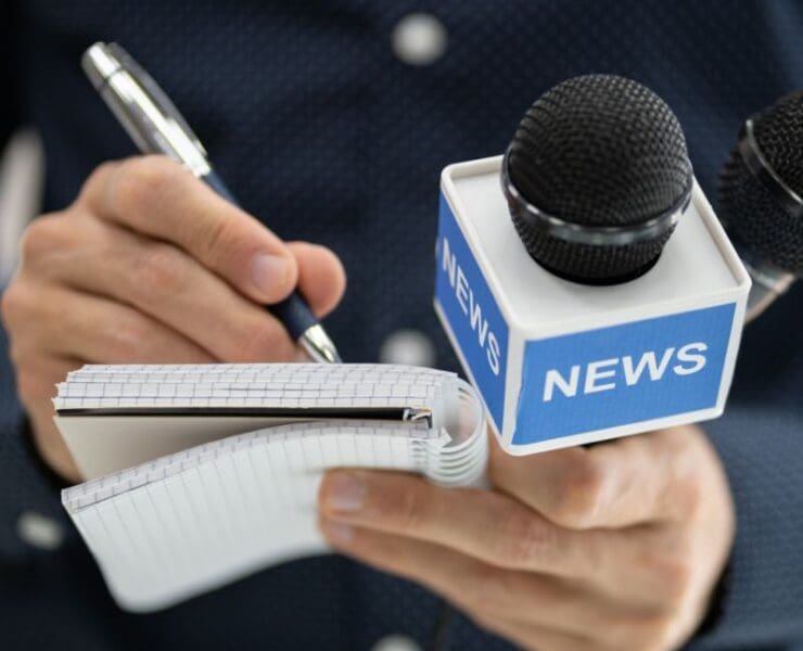 Close-up of a reporter holding a notebook and news microphones, representing a public appeal for an unidentified person of interest.